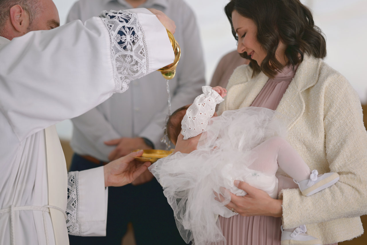 Čin krštenja djevojčice Nike, koju kuma drži u naručju dok ju svećenik polijeva svetom vodicom.