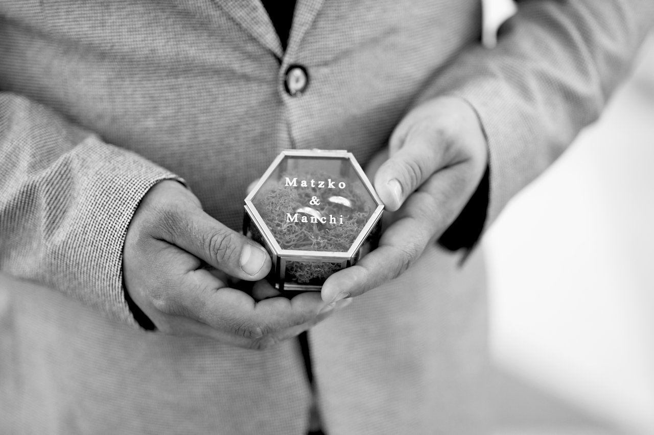 Crnobijela fotografija vjenčanog prstenja u kutijici na kojoj piše Matzko & Manchi, u rukama kuma.