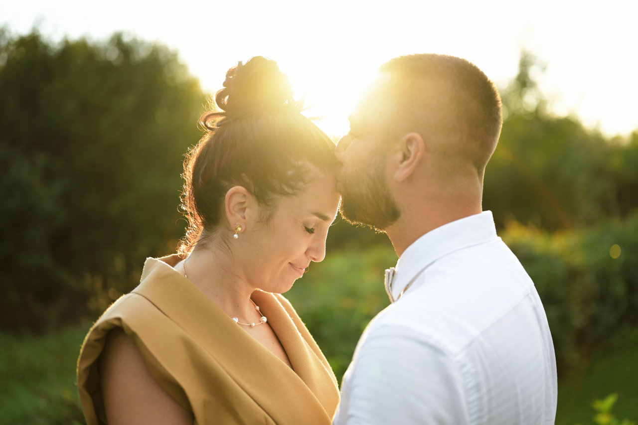 Trenutak nježnosti između mladog para – mladić ljubi djevojku u čelo dok se u pozadini vidi zalazak sunca.