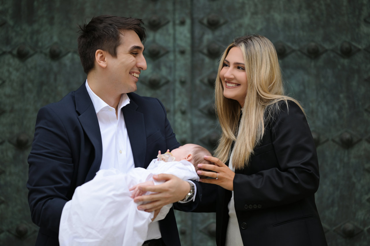 Roditelji se nježno gledaju dok drže bebu ispred ukrasnih vrata crkve; emotivna fotografija krštenja u Zagrebu.