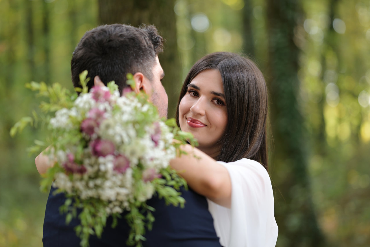 Nježno nasmiješena mladenka grli mladoženju i gleda u objektiv.