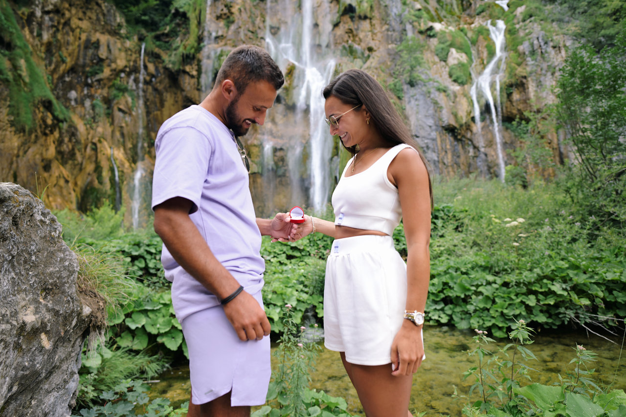 Fotografiranje zaruka na Plitvicama – mladić i djevojka se nasmiješeni dive zaručničkom prstenu dok se u pozadini vidi veliki slap.