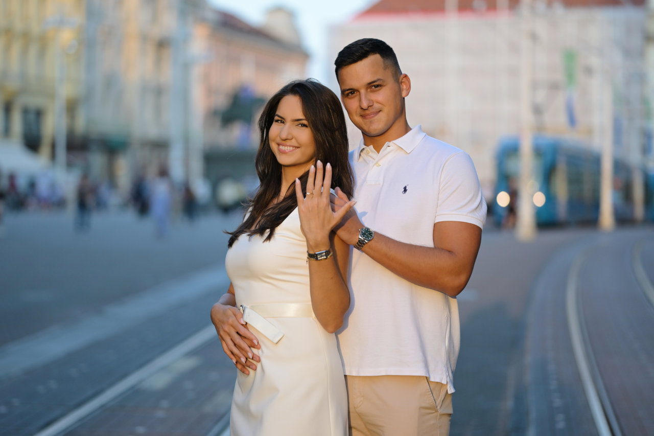 Nasmijani par na Trgu bana Jelačića u Zagrebu: djevojka pokazuje zaručnički prsten, u pozadini plavi tramvaj i arhitektura Zagreba.