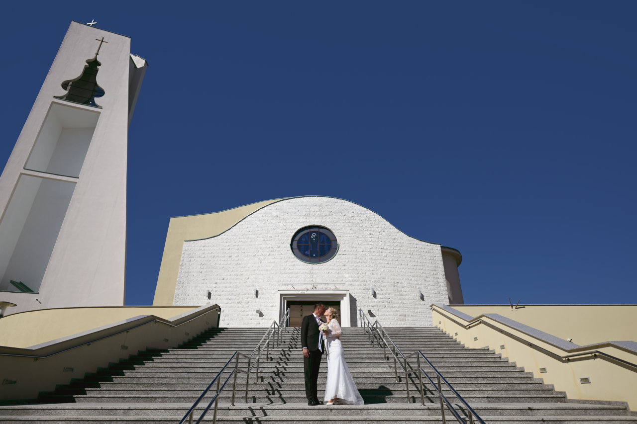 Vjenčani par na stepenicama crkve u Zagrebu, uz modernu arhitekturu i plavetnilo neba.