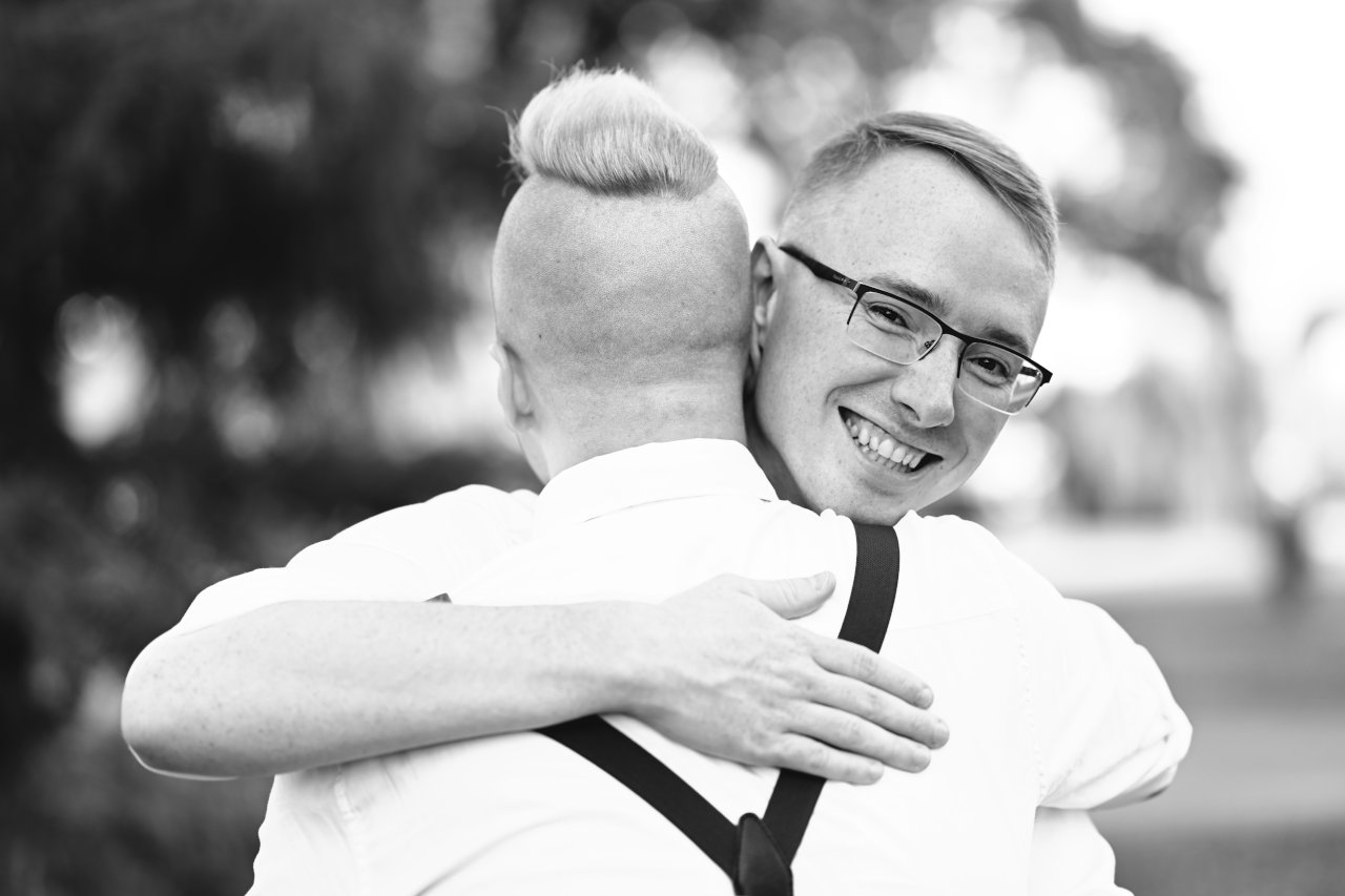Crnobijeli krupni kadar nasmiješenog muškarca s naočalama koji grli svojeg partnera.