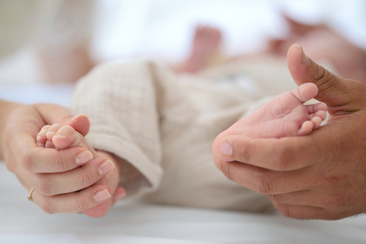 Roditeljske ruke nježno drže sitna stopala bebe u krupnom planu na bijeloj podlozi.