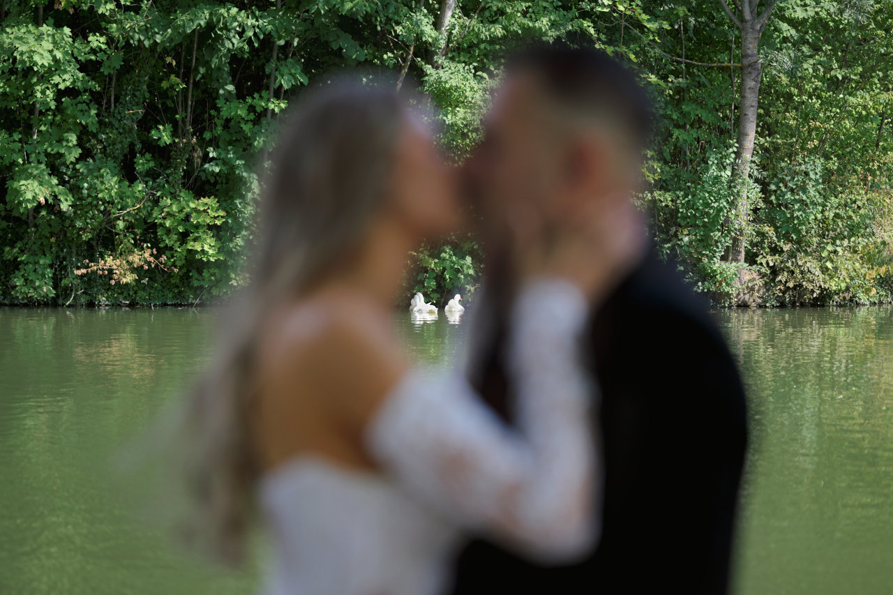 Mladenci u poljupcu izvan fokusa uz jezero Granešina u zagrebačkoj Dubravi, s dva bijela labuda u fokusu na jezeru.