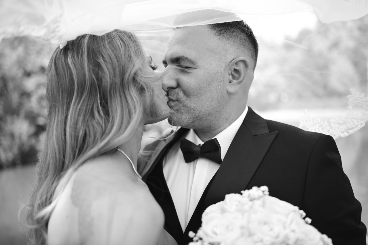 Intimni crnobijeli portret mladenke i mladoženje u odijelu s leptir mašnom kako se ljube ispod vela.